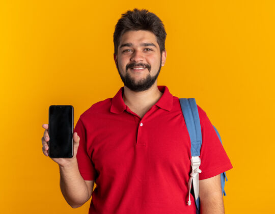 积极年轻的留着胡子的学生 穿着红色马球衫 背着背包 拿着智能手机 微笑着 自信 快乐 积极的站着小伙子红色胡子