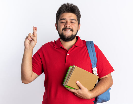 站着年轻的留着胡子的学生 穿着红色马球衫 背着背包 拿着笔记本 手交叉着手指 站着希望的表情手指背包小伙子
