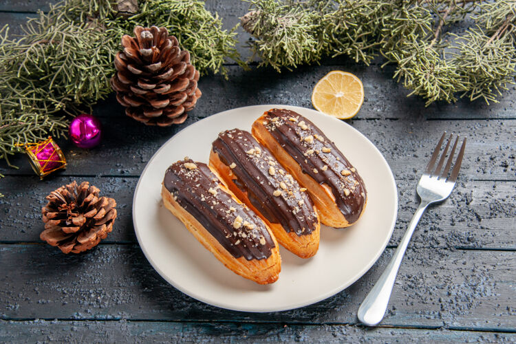 晚餐下图：白色椭圆形圆锥形板上的巧克力蛋糕 冷杉树叶 圣诞玩具 柠檬片和叉子 放在深色木桌上切片叉子圣诞节