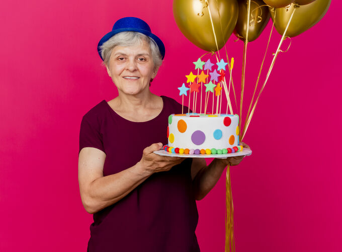 粉色微笑着戴着派对帽的老太太拿着氦气球和生日蛋糕看着粉红色的相机穿相机聚会