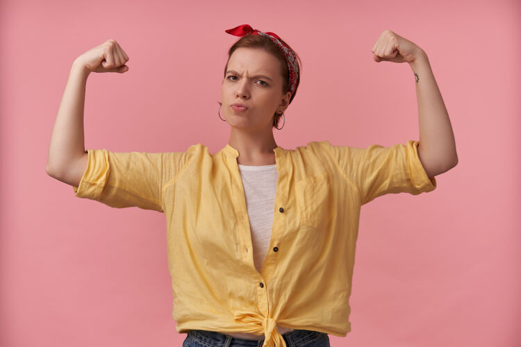 女人身穿黄色衬衫 头戴头巾的强壮的运动型年轻女子站着 双手露出二头肌在粉红色的墙上骄傲能量美丽