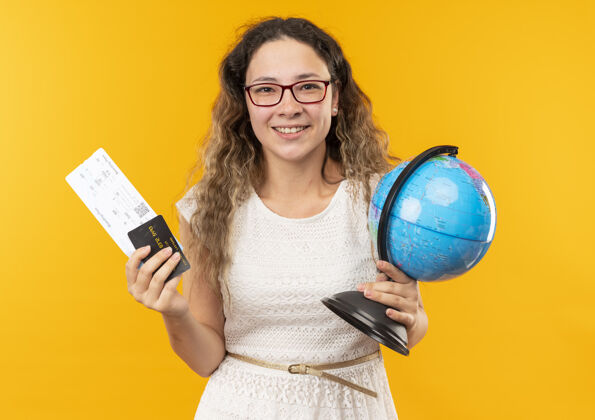 地球微笑的年轻漂亮的女学生戴着眼镜拿着机票 信用卡和地球仪孤立在黄色的墙上票微笑信用