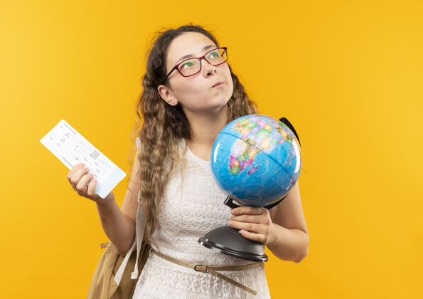 衣服体贴的年轻漂亮的女学生戴着眼镜背着包拿着机票和地球仪孤立地看着黄色的墙上姿势脸飞机