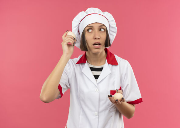 感觉体贴的年轻女厨师 身着厨师制服 手里拿着鸡蛋 看着粉红色墙上孤立的一面厨师思想壁板