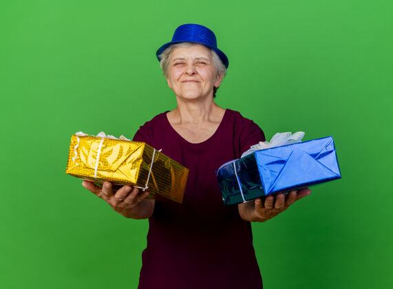 礼物戴派对帽的老太太高兴地拿着礼品盒站在绿色的树上女人聚会帽子
