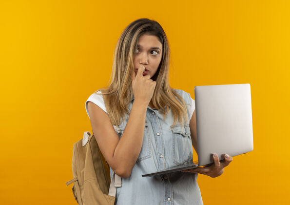 衣服体贴的年轻漂亮的女学生 背着书包 拿着笔记本电脑 手指放在嘴唇上 孤立地看着橙色的墙上姿势女孩人