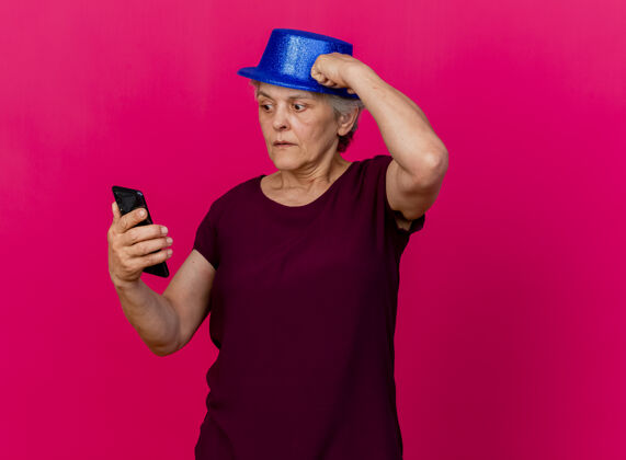 帽子戴着聚会帽的焦虑的老妇人把拳头放在头上看着粉红色的手机焦虑电话聚会