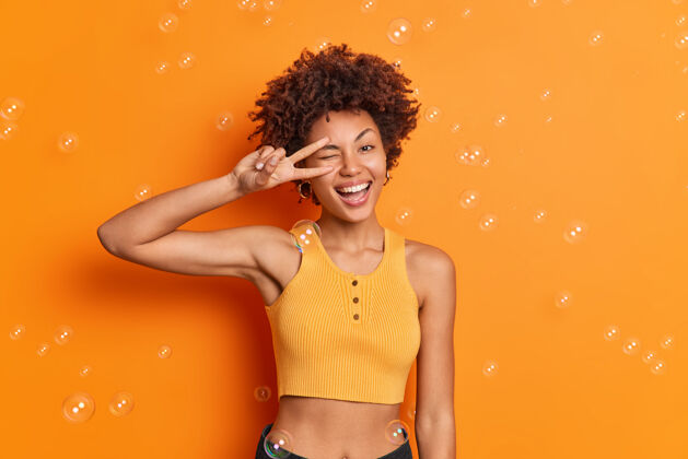 快乐半身镜头 一头卷发的美国黑人女性 身材匀称 穿着不规则剪裁的上衣 眼神中流露出和平的姿态 笑容在橙色的墙上被广泛隔离女人休闲表演