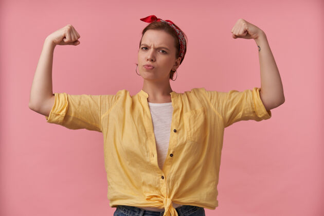 女人身穿黄色衬衫 头戴头巾的强壮的运动型年轻女子站着 双手露出二头肌在粉红色的墙上骄傲能量美丽