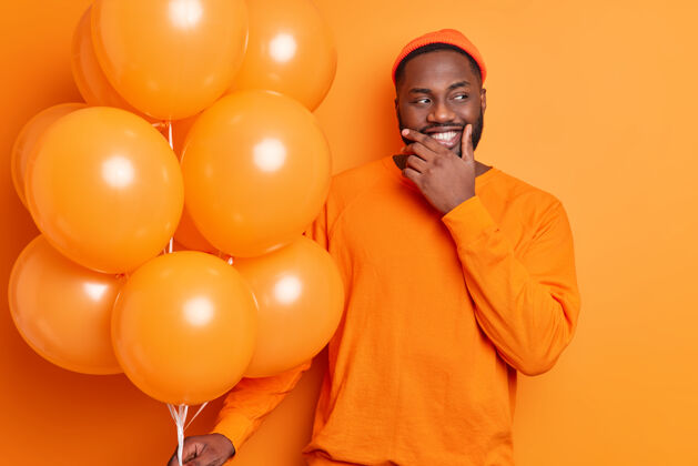 聚会积极的胡须男人拿着一堆气球庆祝节日穿着休闲套头衫和帽子隔离在橙色的墙上参加派对美丽生日明亮