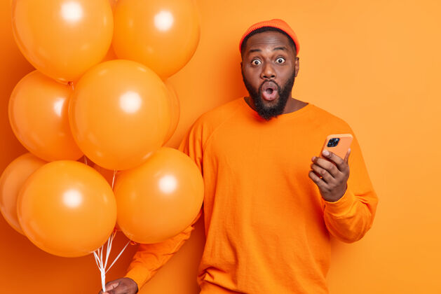 成人水平拍摄的震惊的胡须男子惊讶地盯着相机穿着随意举行手机和一堆气球是在聚会隔离橙色墙担心紧张脸