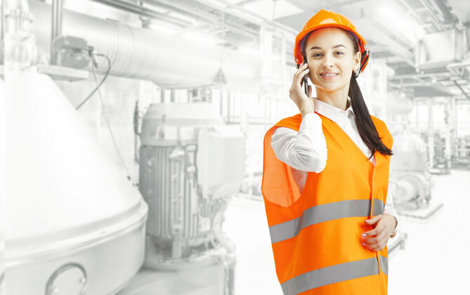 建筑戴着橙色头盔的女建筑工人手持手机站在工业墙上平等建筑商安全帽