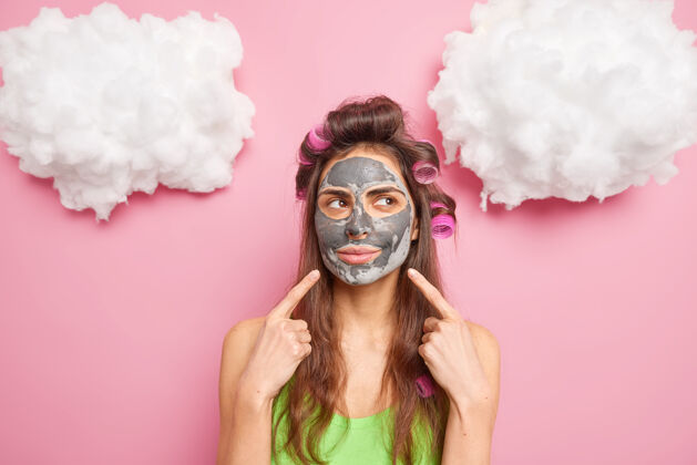 室内欧洲女人指着脸示范美容产品敷在脸上让卷发集中在一边隔离在玫瑰色的墙上面部脸欧洲