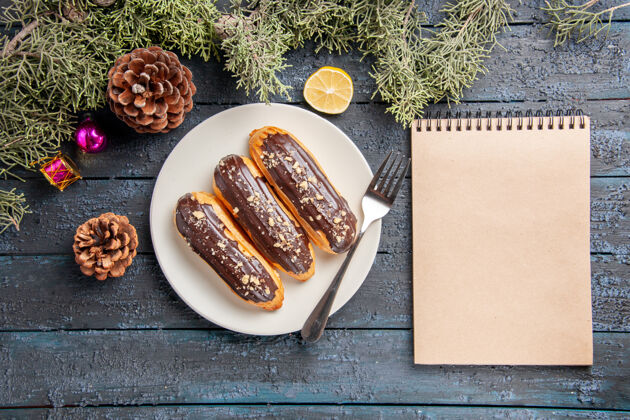 圣诞节俯瞰图：白色椭圆形圆锥体上的巧克力蛋糕和叉子冷杉树叶圣诞玩具柠檬片和一本笔记本放在深色木桌上叶子午餐烹饪