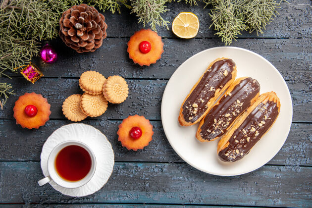 树枝俯视图白色椭圆形平板上的巧克力蛋糕冷杉树枝圣诞玩具柠檬片一杯茶饼干和纸杯蛋糕在深色木质地面上茶膳食饼干