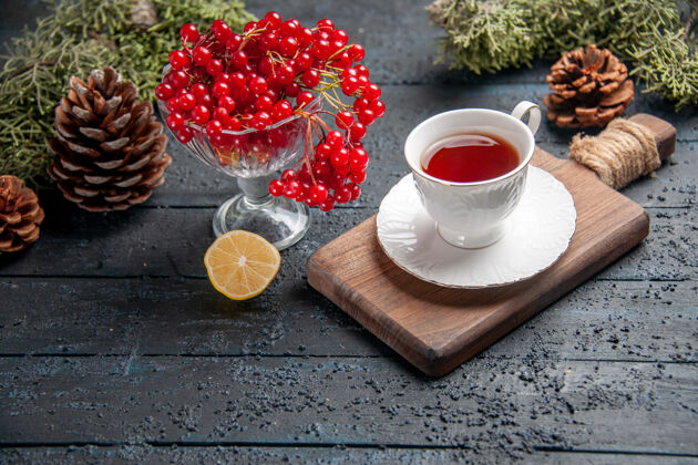 食物下图：一杯红醋栗 一杯茶放在砧板上 一片柠檬松果放在深色木质背景上早餐柠檬茶