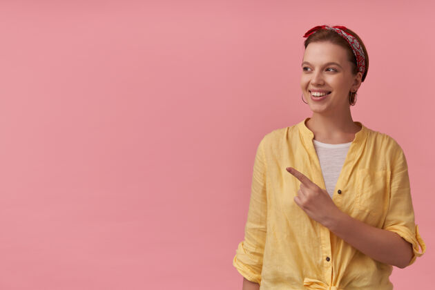 头带穿着黄色衬衫 头上戴着头巾的快乐迷人的年轻女子看着粉色墙上的广告空间 并指着一边指向美丽空
