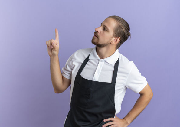 人体贴的年轻英俊的理发师穿着制服 把手放在腰上 抬起手指看着紫色墙上孤立的一面理发师提高手指