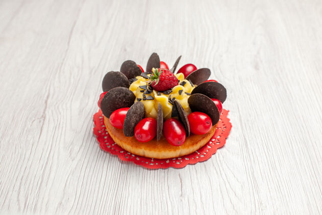 磨碎下图：白色木质背景上的红色椭圆形蕾丝小饰物上有浆果巧克力蛋糕盘子水果食品