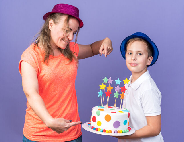 请戴着蓝色派对帽的年轻斯拉夫男孩高兴地拿着生日蛋糕 母亲戴着紫色派对帽站在那里 指着隔离在紫色墙上的蛋糕 留着复制空间帽子孤立的蛋糕