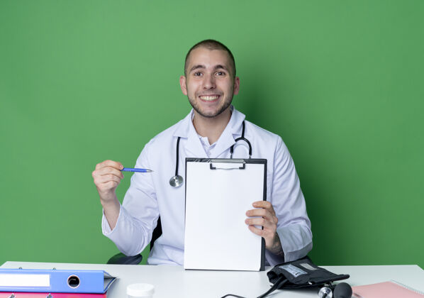 医生面带微笑的年轻男医生穿着医用长袍和听诊器坐在办公桌旁 拿着工作工具 拿着笔指着隔离在绿色墙上的剪贴板抱着绿色脸