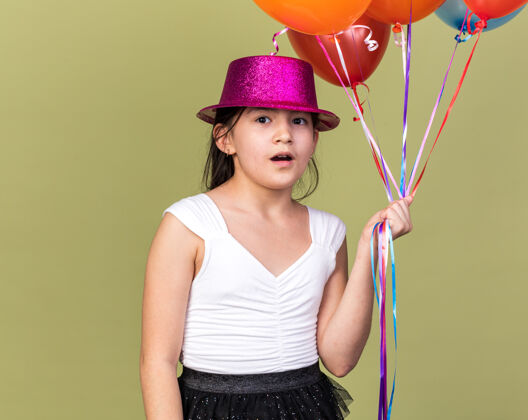 白种人惊讶的年轻白人女孩 戴着紫色的派对帽 手里拿着氦气球 孤立地放在橄榄绿的墙上 留着复制空间女孩抱着惊喜