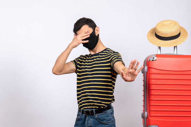 视图正面图：戴着黑色面具的年轻游客站在红色手提箱旁 用手闭上眼睛红色手面具