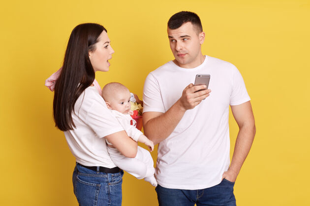 可爱愤怒的年轻母亲手里抱着婴儿 看着丈夫在用手机天真童年健康