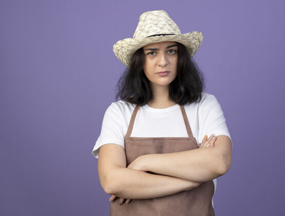 帽子自信的年轻黑发女园丁身着制服 戴着园艺帽 双臂交叉 孤零零地站在紫色的墙上市民花园制服