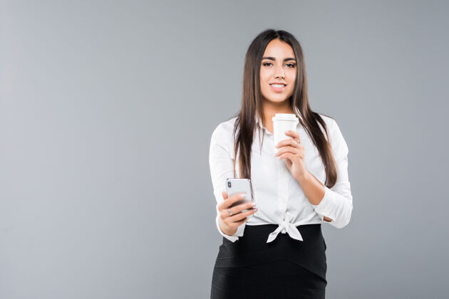 杯子一个满意的年轻女商人的肖像 一边用手机一边拿着一杯咖啡走在白色的隔离成人交流电话