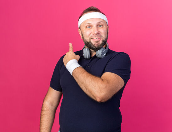 腕带令人高兴的成年斯拉夫运动男子戴着耳机 戴着头带和手带 指向后面的粉红色墙壁与复制空间隔离男人请孤立型