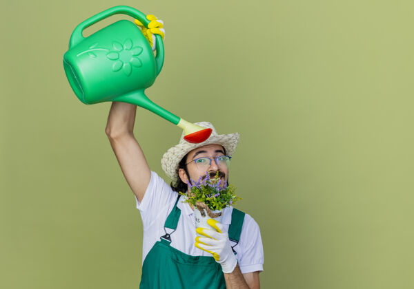 年轻人年轻的留着胡须的园丁 穿着连体衣 戴着帽子 手里拿着浇灌罐和盆栽浇灌植物 站在淡绿色的墙上人站着人