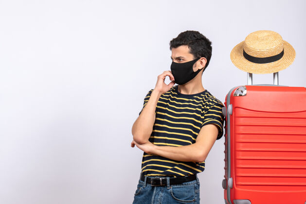 视图正面图持怀疑态度的年轻游客戴着黑色面具站在红色手提箱附近面具男性帽子