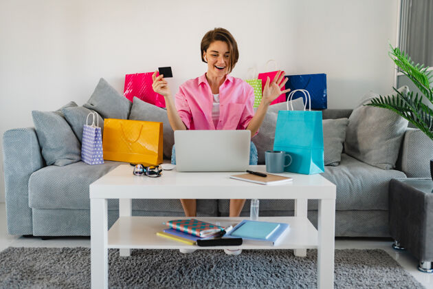 在线在家里的沙发上 穿着粉色衬衫的快乐微笑的女人在五颜六色的购物袋中拿着信用卡 在笔记本电脑上在线支付包支付订单