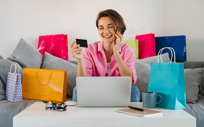 销售在家里的沙发上 穿着粉色衬衫的快乐微笑的女人在五颜六色的购物袋中拿着信用卡 在笔记本电脑上在线支付设备女人技术