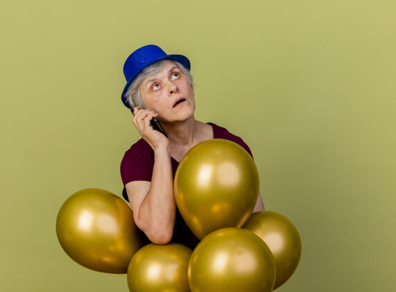 氦戴着派对帽的震惊的老妇人拿着氦气球打电话 孤零零地看着橄榄绿的墙上女人电话老人