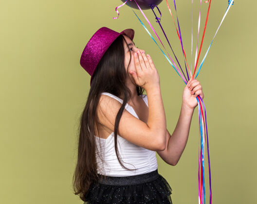 孤立惊讶的年轻白人女孩 戴着紫色派对帽 手放在脸上 看着橄榄绿墙上的氦气球 还有复制空间白种人女孩举行