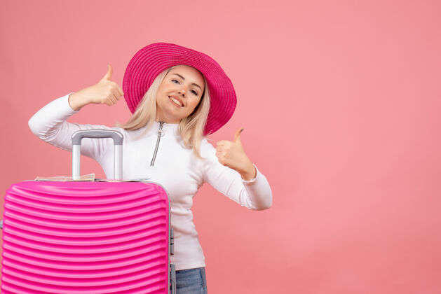 赠送正面图：站在粉色手提箱后面竖起大拇指的年轻女士快乐年轻女士成人
