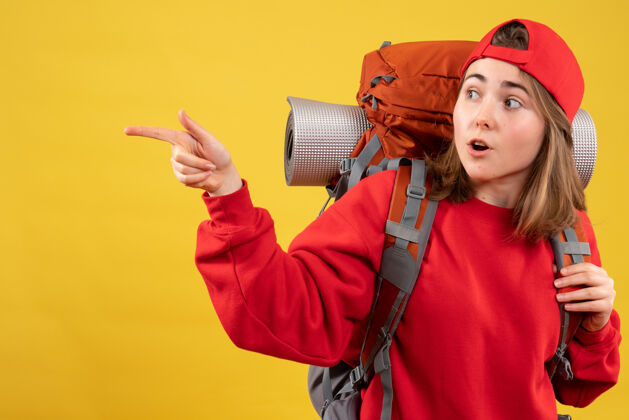 成人正面图：年轻的旅行女孩戴着红色帽子 背包指向左边指帽子背包