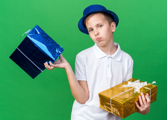 盒子自信的年轻斯拉夫男孩 戴着蓝色派对帽 拿着礼品盒 隔离在绿色墙壁上 有复制空间年轻孤立自信