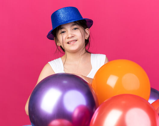 气球微笑着的年轻白人女孩戴着蓝色的聚会帽站在粉红色的墙上 氦气球与复制空间隔离年轻氦女孩