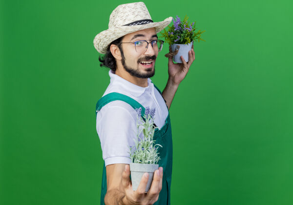 锅年轻的留着胡须的园丁身穿连体衣 戴着帽子 站在绿色的墙上 脸上露出盆栽植物的快乐和积极的微笑人微笑市民