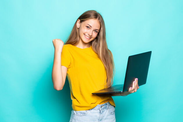 白色在绿松石墙上 一个年轻的女人正在用笔记本电脑摆姿势电脑女孩成功