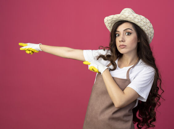 脸印象深刻的年轻女园丁穿着制服戴着园艺帽和手套点在一边隔离在粉红色的墙上人帽子姿势