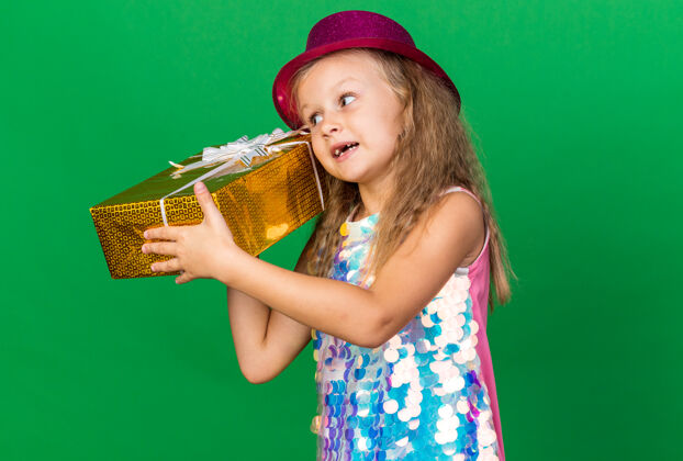 孤立可爱的金发小女孩 戴着紫色派对帽 耳边放着礼品盒 隔着绿色的墙壁 留着复印空间抱着生日帽子