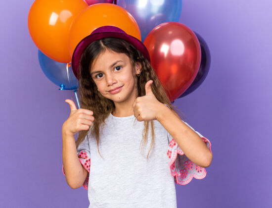 女孩可爱的白人小女孩 戴着紫色派对帽 竖起大拇指站在氦气球前 氦气球被隔离在紫色的墙上 留着复制空间请小氦