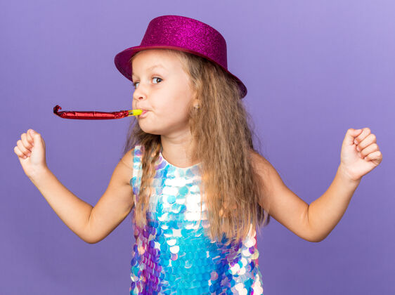 帽子高兴的小金发女孩与紫罗兰党帽子吹党哨子 并保持拳头孤立在紫色墙上复制空间派对保持生日