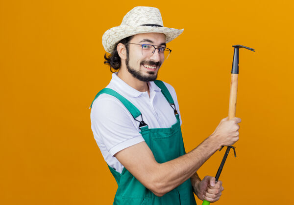 脸年轻的留着胡子的园丁 穿着连体衣 戴着帽子 拿着床垫和小耙子 站在橙色的墙上微笑着看着前面姿势人年轻人