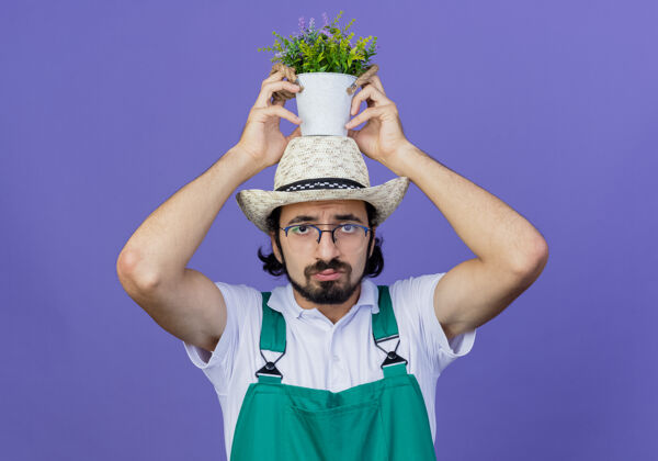 表情年轻的留着胡子的园丁 穿着连体衣 戴着帽子 头上顶着盆栽植物 站在蓝色的墙上 带着悲伤的表情看着前方年轻脸感觉