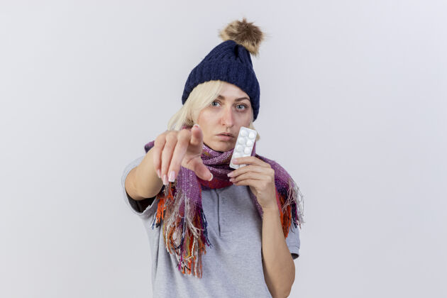 表情自信的年轻金发病女人戴着冬天的帽子和围巾 手里拿着一包药片 指着隔离在白墙上的前面穿着冬天持有